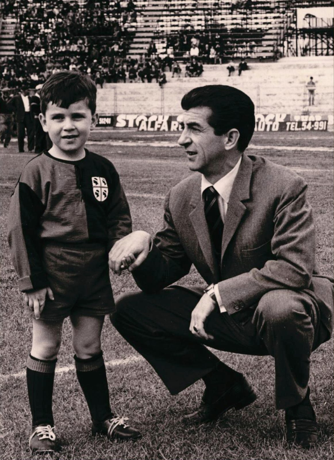 Arrica accovacciato sul prato dello stadio accanto a un bambino con la maglia del Cagliari